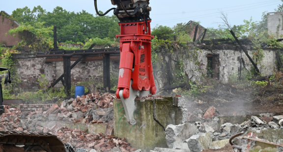 Robot do rozbiórki podnoszący fragment muru w otoczeniu gruzów i zniszczonych budynków.