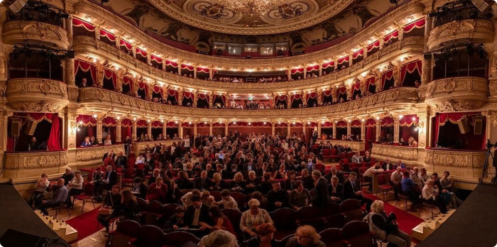 Widok wnętrza eleganckiego teatru z zajętymi miejscami na widowni, w złotych i czerwonych wykończeniach, z tłumem widzów oczekujących na rozpoczęcie przedstawienia.